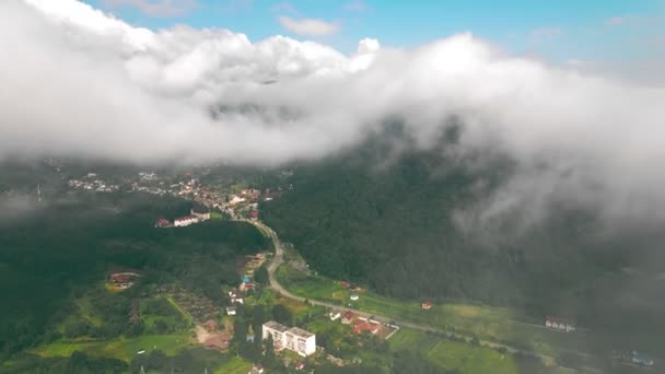 Luftaufnahme Flug über die Wolken, durch die man das Dorf zwischen den Bergen sehen kann. Blick von oben auf die Wolken, die die Berge und Siedlungen in den Bergen bedecken. — Stockvideo