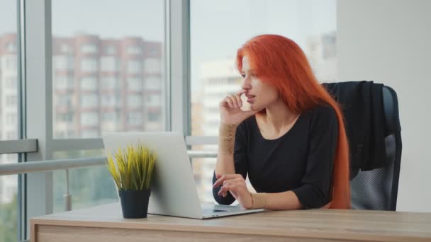 Joven mujer atractiva de aspecto caucásico y cabello rojo trabajando en la oficina en un ordenador portátil. La mujer aguanta la respiración, piensa y trabaja. — Vídeos de Stock