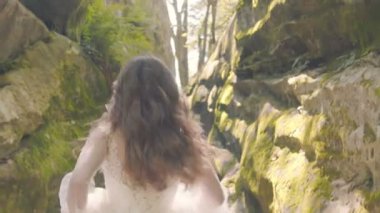 Gelinlik giymiş gelin koşar ve kameraya gülümser. Arka plandaki güneş ışınları flaşlarla kamerayı aydınlatıyor. Gelin kayaların arasında koşar