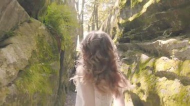Gelinlik giymiş gelin koşar ve kameraya gülümser. Arka plandaki güneş ışınları flaşlarla kamerayı aydınlatıyor. Gelin kayaların arasında koşar