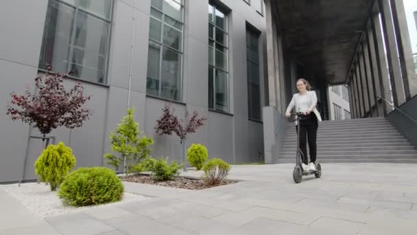 Junge erfolgreiche Frau fährt an einem sonnigen Tag einen Elektroroller und genießt das Leben vor dem Hintergrund schöner Grünanlagen im Technologiepark. — Stockvideo