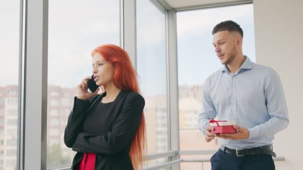 Un hombre le da un regalo a una mujer de pelo rojo. Una mujer que habla por teléfono recibe un regalo de su marido y se alegra por el regalo recibido en la oficina. — Vídeos de Stock
