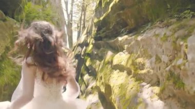 Gelinlik giymiş gelin koşar ve kameraya gülümser. Arka plandaki güneş ışınları flaşlarla kamerayı aydınlatıyor. Gelin kayaların arasında koşar
