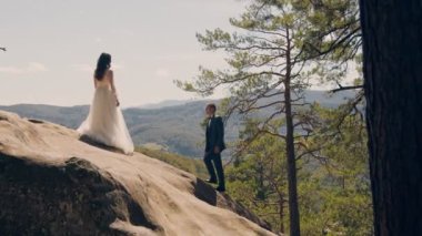 Bir çift mutlu gelin sırtı kameraya dönük olarak dağda duruyor ve ağaçlarla kaplı dağlara bakıyor. Birbirine aşık genç bir çiftin geleceğine bakıyorum..