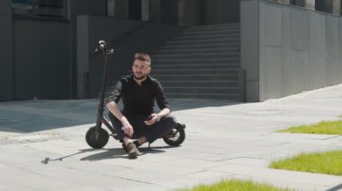 Caddenin ortasında elektrikli scooter 'da oturan ve elinde cep telefonu tutan bir erkek öğrenci. Öğrenci caddede dinleniyor. Elektrikli gözlüklü beyaz adam.