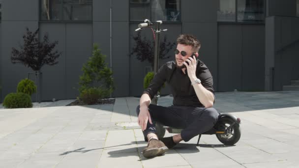 Студент-мужчина садится на электрический скутер посреди улицы и общается по мобильному телефону, показывая негативные эмоции. Белый мужчина в черных очках на электроскутере. — стоковое видео