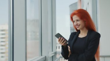 Cep telefonundan mesaj alan ve mutlu bir şekilde gülümseyen genç ve mutlu bir kadın. İş yerindeki bir kadının olumlu duyguları. Kızıl saçlı ve pencerenin yanında telefonu olan bir kızın portresi..
