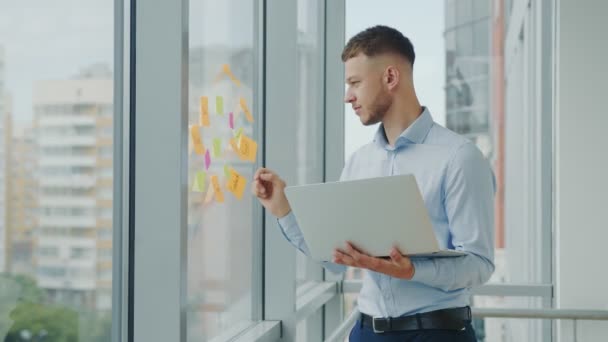 Jovem empresário bem sucedido no escritório pela janela trabalhando com um laptop. O homem resolve tarefas estratégicas de negócios. — Vídeo de Stock