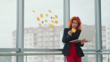 Laptop ve çıkartmalarla ofis işi. Genç kızıl saçlı kadın elinde bir dizüstü bilgisayar tutuyor ve pencereden bir çıkartma alıyor ve onu inceliyor. İş yerindeki iş kadını..