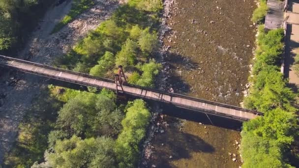 Вид с воздуха со Старого деревянного подвесного моста через горную реку. Девушка стоит на деревянном мосту и смотрит в будущее. Карательный пейзаж горной реки в солнечный день. — стоковое видео