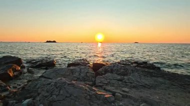 Adriyatik Denizi üzerinde altın gün batımı önplanda kayalık kıyı şeridi, dalgalara ve ufka yansıyan ılık güneş ışığı sakin doğal deniz manzarası atmosferi yaratıyor.