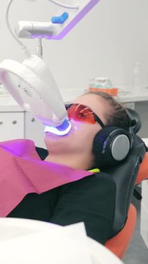 LED beyazlatma sırasında koruyucu gözlük takan hasta klinikte bir sandalyede oturuyor. Modern bir diş kliniğinde dişlerin foto beyazlatılması.