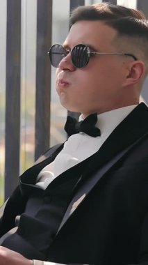 Smokinli ve siyah gözlüklü bir adam bir bardak alkol ve sigarayla balkonda oturuyor. Genç bir adam sigarasını içti ve kameraya duman üfledi ve bir bardak viskiden içti..