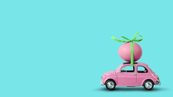 Pinkfarbenes Spielzeugauto mit einem Osterei auf dem Dach auf mintblauem Hintergrund. — Stockfoto