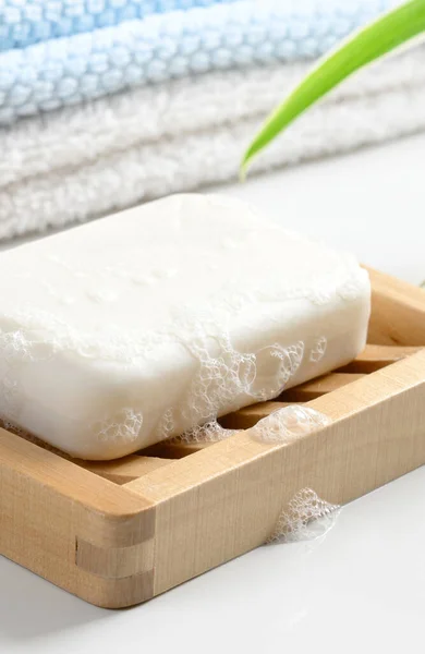 White Soap bar with foam on wooden soap dish and cotton towels on white table. Closeup