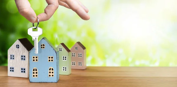 Hand with house key and miniature houses on green nature background