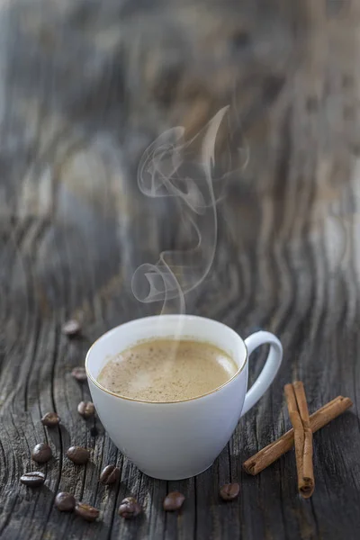 White steamy cup near cinnamon sticks in coffee beans, on old wooden table — Stock Photo, Image