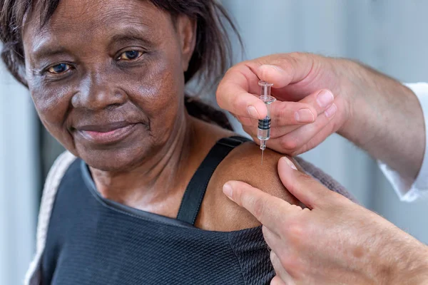 Afrykańska starsza kobieta otrzymująca szczepionkę przeciw chorobie, kaukaskie ręce trzymające strzykawkę z igłą obok górnej części ramienia — Zdjęcie stockowe