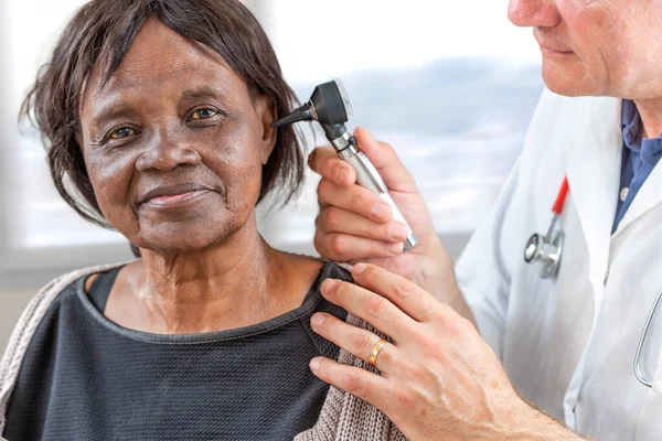Doctor sosteniendo otoscopio y examinando la oreja de una mujer africana mayor . — Foto de Stock