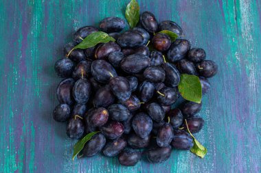 Canlı bir taze erik yığını eskimiş ahşap bir yüzeyde duruyor, koyu mor renklerini ve yeşil yapraklarını sergiliyorlar. Düzenleme meyvenin doğal güzelliğini vurguluyor..