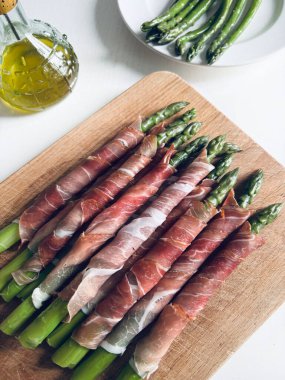 Tahta kesme tahtasına sarılmış jambonlu taze kuşkonmaz. Tam üstte.