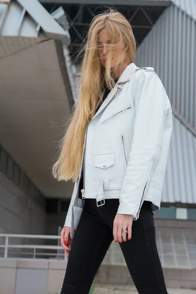 Blonde woman in the urban background. Beautiful young girl wearing a white leather jacket standing in the street.