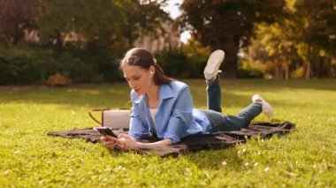 Genç bir kadın park çimlerinde battaniyeye uzanıp kulaklıkla müzik dinliyor, telefon kullanıyor, bacaklarını sallıyor, dışarıda dinleniyor. Mutlu yaşam tarzı, boş zaman, yaz havası, kaygısız, huzurlu an..