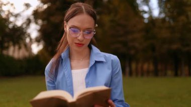Genç bir kadın parkta kitap okuyor, sonra kapatıyor ve uzaktan tatmin olmuş görünüyor. Rahat öğrenci yaşam tarzı, açık hava eğlencesi, sakin doğa, mutluluk, özgürlük, huzurlu yaz titreşimleri.