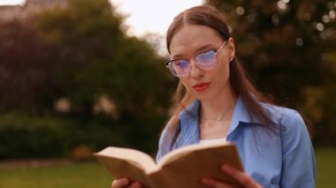 Gözlüklü genç kız parkta ya da kampüste çimenlerde oturur, dikkatle kitap okur, sayfaları çevirir, odaklanır ve rahatlar, dışarıda huzurlu çalışma anlarından zevk alır..