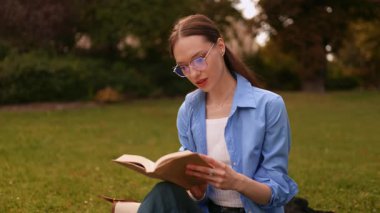 Gözlüklü gülümseyen genç kız parkta ya da kampüste çimlerle ilgili bir kitap okuyor, arkadaşlarına el sallıyor, dışarıda çalışmaktan ve rahatlatıcı anlardan keyif alıyor..