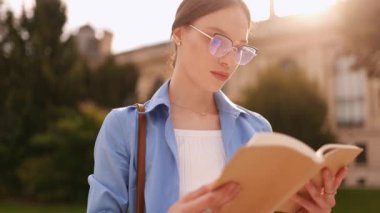 Üniversite kampüsünün yanında gözlük okuyan, gün ışığında gülümseyen, öğrenme, neşeli yaşam tarzı, eğitim, bilgi ve açık havaya odaklanmış mutlu genç bayan öğrenci..