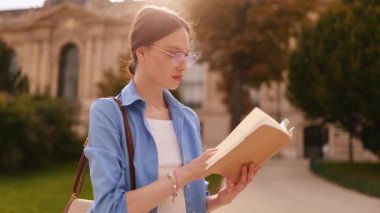Üniversite kampüsünün yanında duran gözlüklü genç bir bayan, ilginç bir şekilde kitap okuyor, eğitime odaklanıyor, bilgi, yaşam tarzı, öğrenim anı.