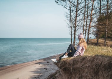 Pembe şapkalı genç bir kadın kayalıklarda tek başına oturuyor, deniz, özgürlük konsepti, huzurlu atmosfer, meditasyon, fotokopi alanı, Letonya, Jurkalne