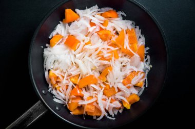 Siyah bir arka planda bir kızartma tavasında bitkisel yağda doğranmış çiğ soğan ve taze portakal biber, üst görünüm