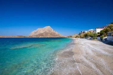 Yunanistan 'ın Telendos adasında, açık mavi gökyüzünün altında turkuaz su ve dağlarla kaplı bir sahil.
