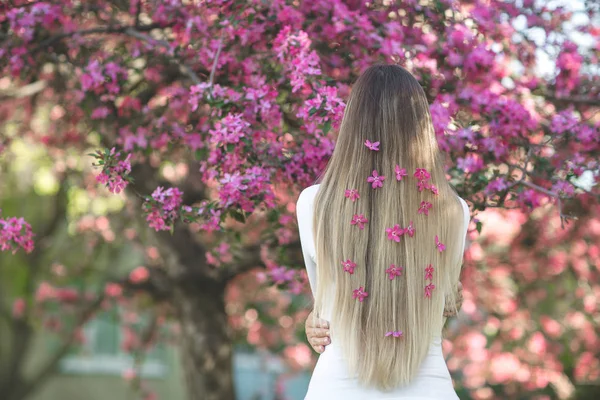 Unrecognizable woman standing back side to camera with long blond hair with flowers in her hair. Female on spring background. Lady outdoors.