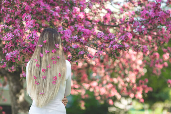 Unrecognizable woman standing back side to camera with long blond hair with flowers in her hair. Female on spring background. Lady outdoors.