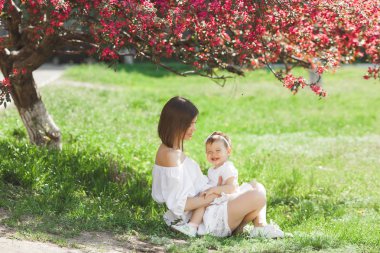 Küçük bebek kızı ile genç güzel annesinin portresi. Yine de sevgi dolu aile, yakın. Çocuğunu pembe çiçekler içinde tutarak ve gülümseyen çekici kadın.