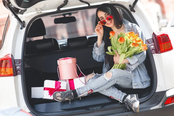 Young attractive woman in the car with presents box, gift and flowers. Beautiful lady in spring time with bouquet of tulips. Female in the automobile.