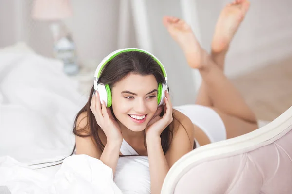 Young attractive woman listenning to the music. Attractive girl in the headphones. Woman indoors in the bed relaxing. Lady wearing earphones.