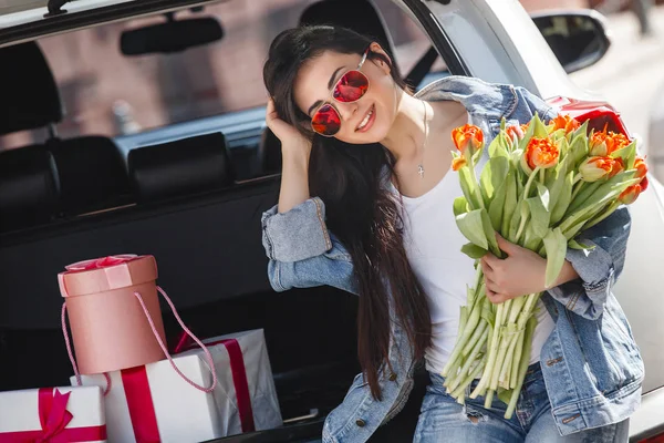 Young attractive woman in the car with presents box, gift and flowers. Beautiful lady in spring time with bouquet of tulips. Female in the automobile.