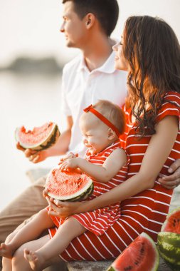 Mutlu genç aile köprüde suyun yanında oturuyor. Anne, baba ve küçük kızları birlikte oturuyorlar. Mutlu bir aile dışarıda. Gülümseyen insanlar.