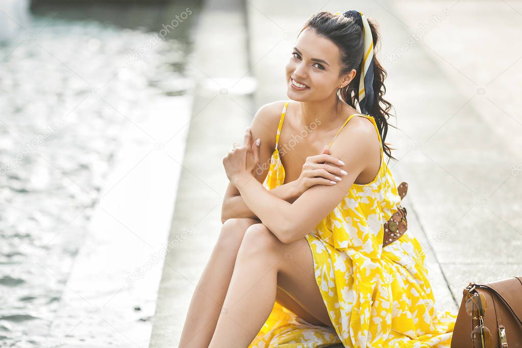 Beautiful woman sitting near the fountain. Lady in yellow dress. Stylish woman. Female outdoors. Trendy adult woman with kerchief in the hair.