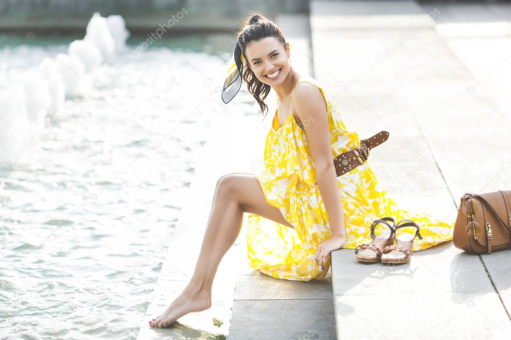 Beautiful woman sitting near the fountain. Lady in yellow dress. Stylish woman. Female outdoors. Trendy adult woman with kerchief in the hair.