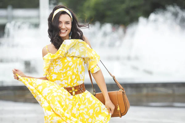 Attractive young woman walking the city. Lady in yellow long dress near the fountain. Beautiful female on summer background.