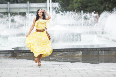 Fıskiyenin yanında çekici genç bir kadın. Güzel kadın açık havada. Sarı elbiseli bayan portresi.
