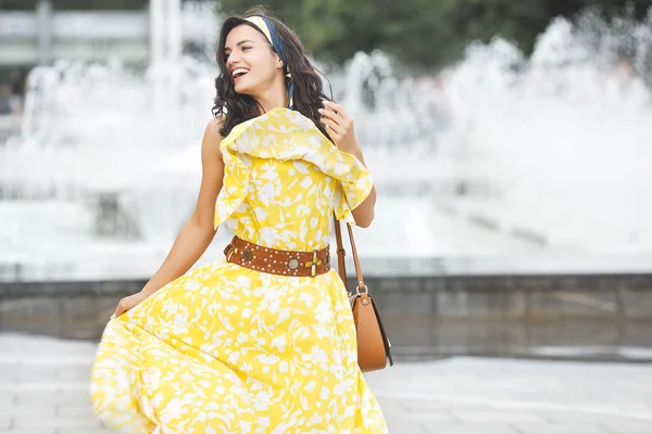 Attractive young woman near the fountain. Beautiful female outdoors. Lady in yellow dress portrait.