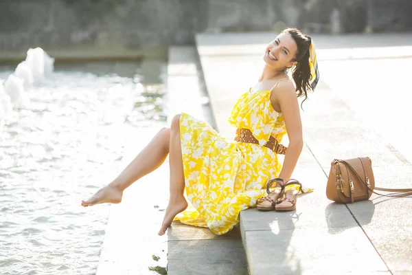Attractive young woman near the fountain. Beautiful female outdoors. Lady in yellow dress portrait.