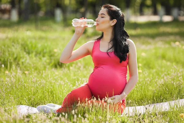 若い妊婦は屋外でフィットネスエクササイズをし きれいなボトル入り飲料水を飲んでいます 新鮮な空気の上でヨガをしている女性を期待して — ストック写真