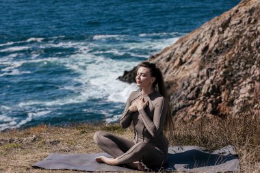 Kadın pratisyen, meditasyon yapar ve doğal güzelliklerle çevrili okyanusun kenarındaki yoga minderine uzanır. Rahatlamayı ve iç huzuru teşvik eder.